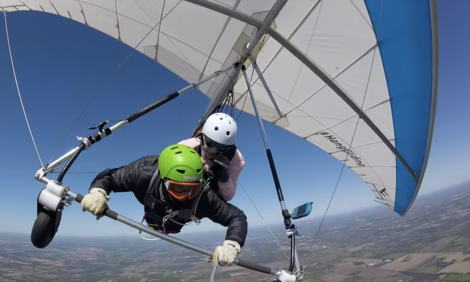 Two people hang gliding high above the ground, with one piloting and the other observing, against a clear blue sky