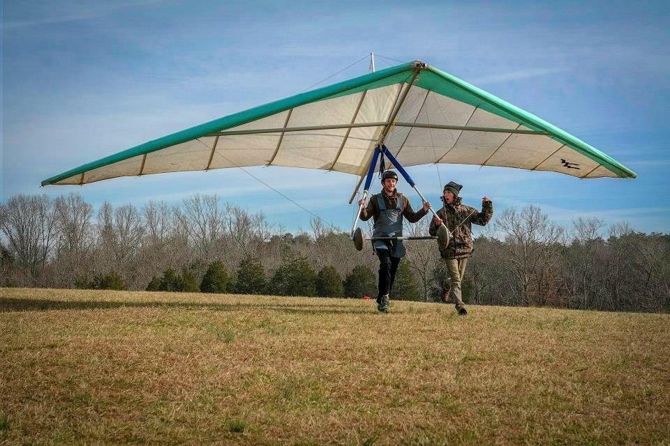 A man on a hang glider runs across a field next to a girl