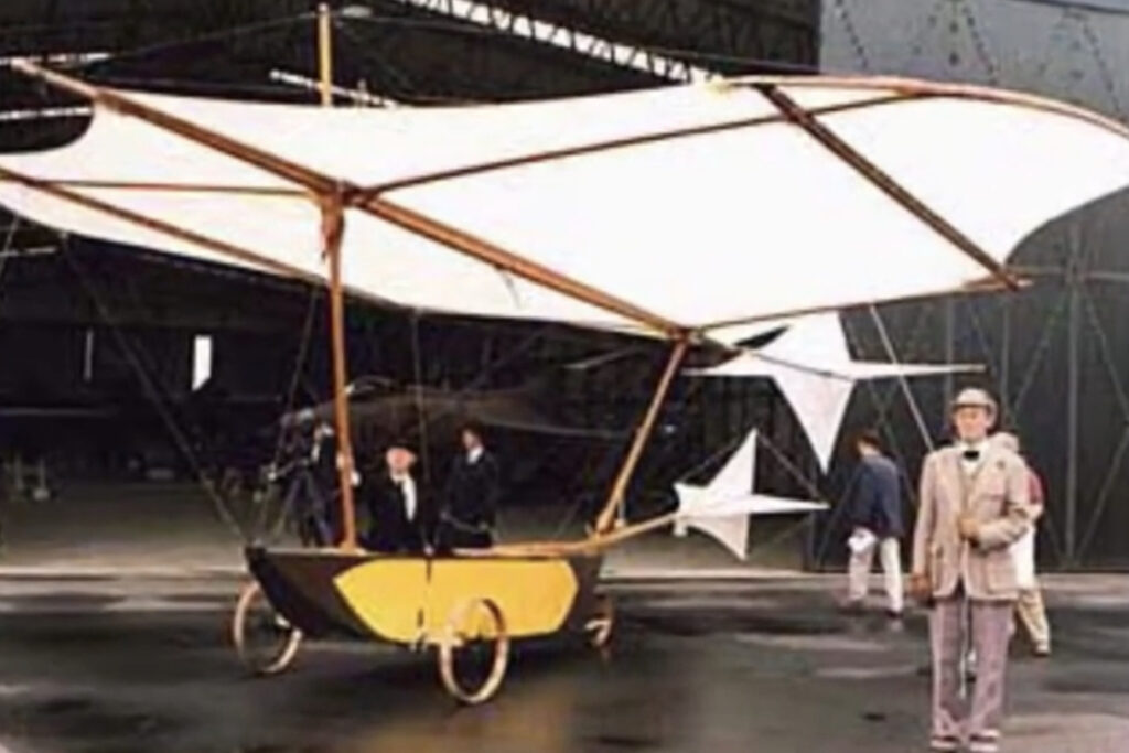 an early model hang glider with a large wing structure, with people around in an air hangar