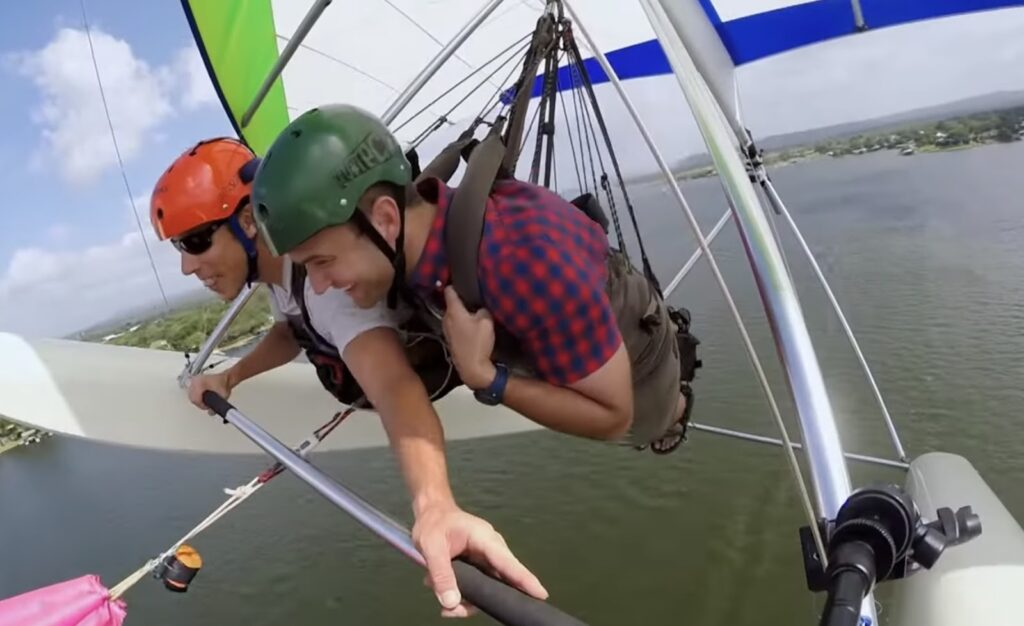 Two hang gliders in mid-air above a lake, with one pilot controlling the glider and the other enjoying the ride