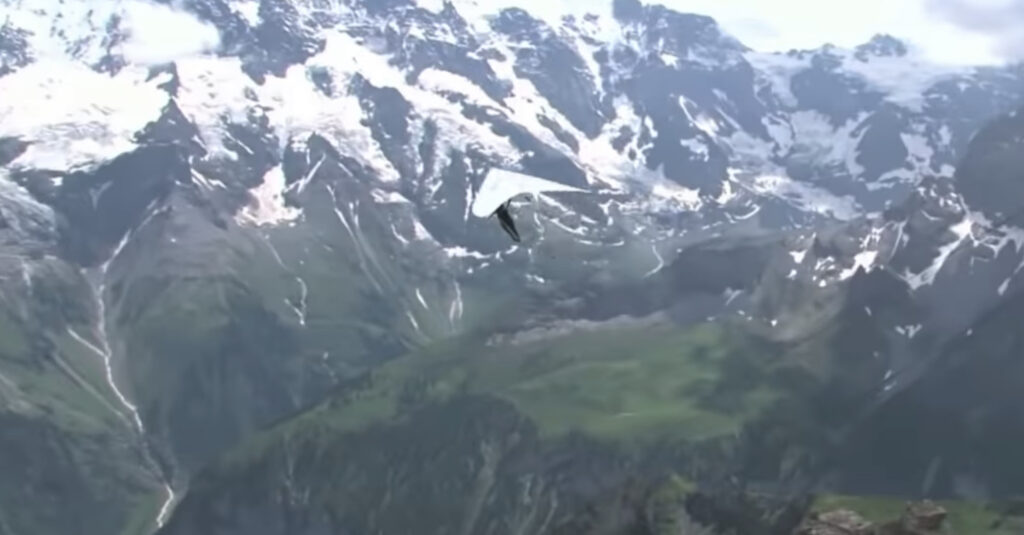 A hang glider flying over rugged mountainous terrain with patches of snow