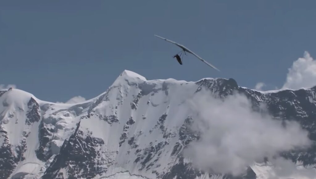 A hang glider soaring in the sky with snow-covered mountain peaks and clouds in the background