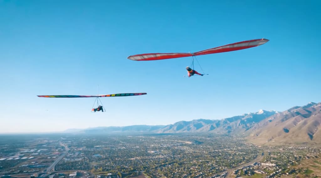 Two hang gliders flying side by side above a suburban area with mountains in the background